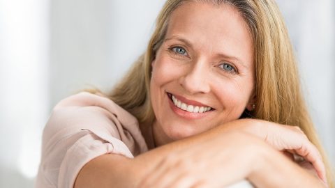 A smiling woman with long blonde hair rests her arms on a surface, looking at the camera.