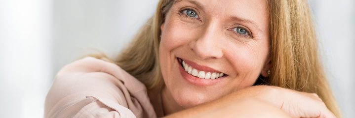 A smiling woman with long blonde hair rests her arms on a surface, looking at the camera.