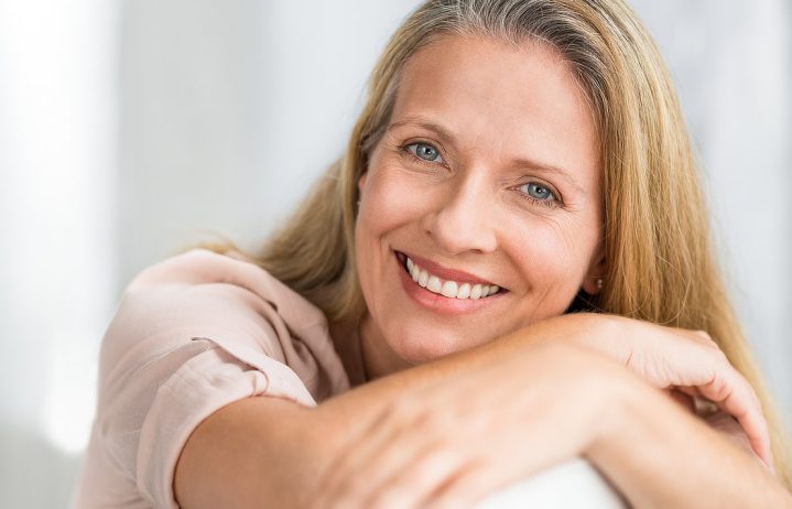 A smiling woman with long blonde hair rests her arms on a surface, looking at the camera.