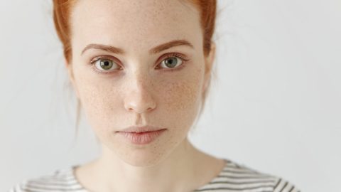 A young woman with red hair, green eyes, and freckles, wearing a white and black striped shirt, looks directly at the camera against a plain light background.