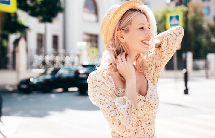 A woman in a light floral dress and straw hat smiles while standing on a sunny city street.